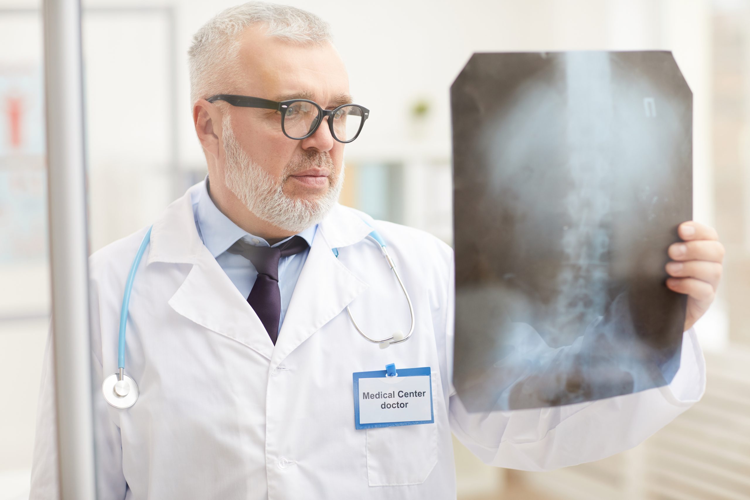 Doctor examining x-ray image Senior doctor in eyeglasses and in white coat holding x-ray image of patient and examining it while standing at hospital